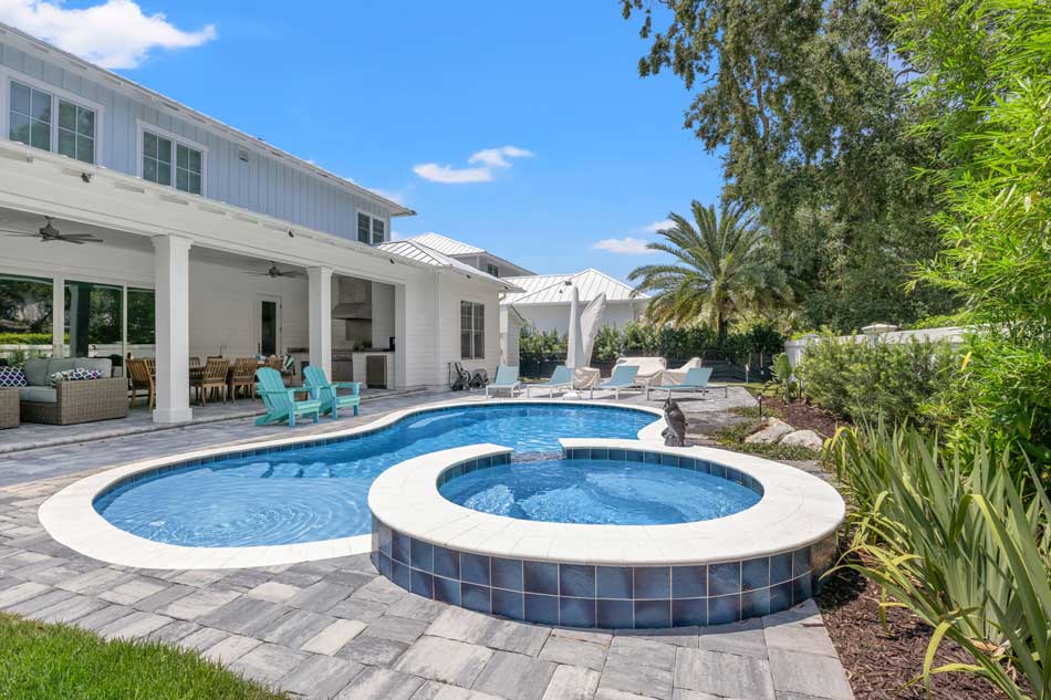 A backyard featuring a modern outdoor space with a kidney-shaped swimming pool and a round hot tub. The area is surrounded by lush greenery and includes a covered patio with furniture, a barbecue area, and palm trees in the background, under a clear blue sky.