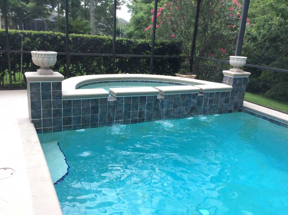 An outdoor swimming pool with a built-in jacuzzi next to it. Both have blue tiles, and they're surrounded by tall greenery and a black fence. The jacuzzi has two large stone planters on either side of its entrance. The water in the pool is clear and blue.