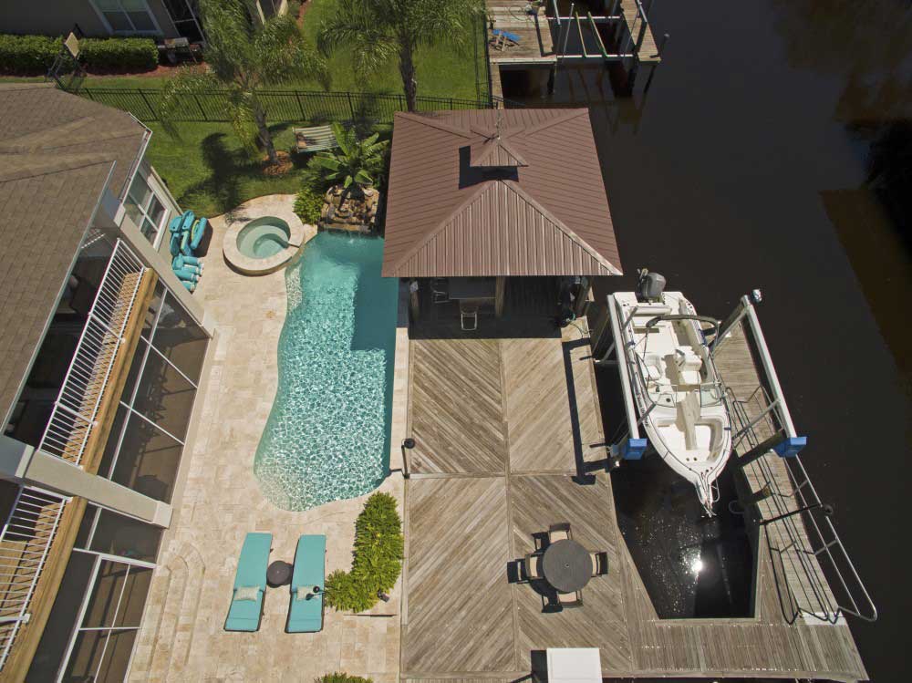 Aerial view of a backyard featuring a pool and jacuzzi surrounded by a stone patio and lounge chairs. Adjacent is a wooden deck leading to a dock with a boat hoist holding a boat above the water. There's a gazebo on the deck and lush greenery in the background.